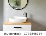 White washbasin with faucet on wooden countertop in minimalist modern bathroom. Scandinavian interior with stylish grey wall and round mirror. Copy space and nobody.