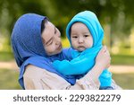 Small photo of Cute young islamic kazakh woman hugging her little crying son, newborn boy. Yappy asian mother in hijab soothes, holding baby in hands outdoors. Family concept