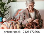 Senior mature woman sitting on sofa at home while knitting and looking at her work in progress. Hobby, retirement, relax concept for elderly smiling female grandmother wearing glasses
