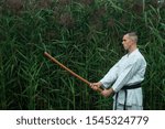 Small photo of male karate in a traditional kimono with a black belt practices karate kick with a bokken. Training takes place against the background of lumps