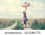 Woman with suitcase standing on road between field of wheat. Backview of girl in hat rising umbrella.