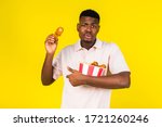 Small photo of African American young guy eats fast food, chicken leg and a whole bucket in his hands. Emotions of condemnation and constraint. Yellow background