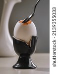 Small photo of White broken egg with a yolk in a broken black ceramic eggcup with a black teaspoon. Shallow depth of field