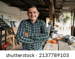 Small photo of Hispanic carpenter smiling proud in wood shop of him - Proud owner of carpentry shop standing with arms crossed