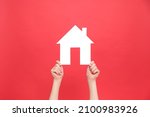 Close up of female hands holding small white paper house, isolated over red color background wall in studio with copy space for promotion content or design. Real estate and family home concept