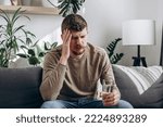 Small photo of Upset young caucasian man suffering from strong headache or migraine sitting on sofa at home with glass of water, guy feeling intoxication and pain touching aching head. Morning after hangover concept
