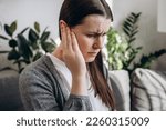 Small photo of Close up portrait of upset young girl holding painful ear, suddenly feeling strong ache. Unhealthy caucasian woman 20s suffering from painful otitis sitting on sofa at home. Health problems concept
