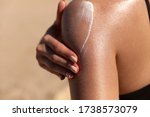 Young woman applying sun cream or sunscreen on her tanned shoulder to protect her skin from the sun. Shot on a sunny day with blurry sand in the background