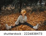 A young happy girl in a fashionable knitted sweater is sitting in the park on autumn orange leaves and covers her face with an autumn bouquet of leaves. Autumn background