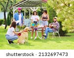 group of friends in the park at a picnic are barbecued, chatting, smiling and playing with a Labrador Retriever dog against the backdrop of greenery and a gazebo