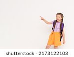 Small photo of A child with a satchel points with his index finger at your advertisement on a white isolated background. Preparatory summer courses for children. Copy space.