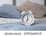 Small photo of Close-up of a round white alarm clock on a table in the bedroom. The hands on the clock show seven o'clock in the morning, time to get up. Retro alarm clock on the table, vintage tone. space for text