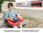 The boy sits on a bench in the school yard and takes out a lunch box from a school backpack. Children have snacks between classes.