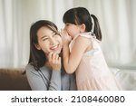 Small photo of Little Asian daughter girl whispering I love you mommy to happy smiling mother in living room