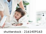 Small photo of Cute Caucasian little toddler baby girl wear bathrobe after bathing is smiling and lying on towel while mother wipe and apply talcum powder on her body at home. Hygiene care for children concept