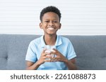 Small photo of Portrait of adorable African Nigerian boy smiling with milk moustache, holding and drinking a glass of milk, sitting on sofa at home, looking at camera. Healthy food and drink for children concept