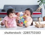 Small photo of Asian young mother and Caucasian 7 months baby newborn girl playing with development toy and bear doll. Single mom lying on floor smiling playing with little daughter, crawling in living room at home