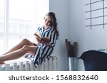 Small photo of Pleasant young lady in trendy dress relaxing next to window and becoming infatuated with cup of tea and book at home office
