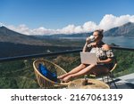 Carefree Swedish woman sitting on hotel balcony with laptop computer while working remote on freelance. tattooed female digital nomad enjoying work on netbook and drink coffee, mountains view