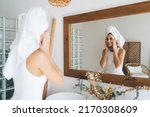 Young beautiful female with towel on head smiling to mirror reflection  standing in the bathroom at home. 30 years old happy woman doing daily morning rituals and cleansing. Enjoying healthy skin care