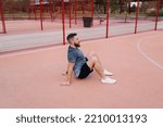 Small photo of Side view of emotionless male athlete in activewear sitting on basketball arena and looking forward while resting after workout on sports ground