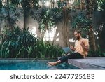 Side view of bearded male in casual clothes sitting on edge of swimming pool with netbook and communicate on video chat. Digital nomad working online on freelance