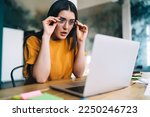 Small photo of Dumbfound young female freelancer wearing eyeglasses looking at netbook screen and sitting with opened mouth at table while working online from home. Studying with online courses or webinar