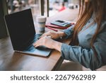 anonymous female freelancer looking at screen while typing on laptop and sitting at table with cup of coffee pen notebook diary smartphone in daytime while attending work for startup in cafe