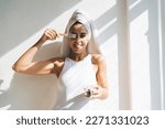 Playful Caucasian lady in white bodysuit and turban applying cosmetic cream with brush and showing tongue while standing near sunlit wall after morning shower