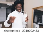 Small photo of Positive African American male in black hat smiling and looking at camera while standing with throwaway cup of coffee in hand in cozy room near mirror with reflection of sofa