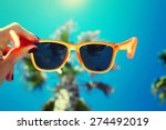 Female hand holding colorful sunglasses against palm tree and blue sunny sky, summer vacation holidays concept, first person shot, looking though glasses, filtered image