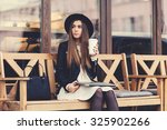 Portrait of a glamorous young woman holding on her knees portable laptop computer while sitting on a wooden bench, stylish female drinking coffee while relaxing after work on net-book during free time