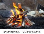 Small photo of Red flames of teepee campfire on dark ground at campsite in wild at overcast, closeup bonfire with firewood on backdrop, autumn relax camping mood, low angle