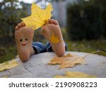 Small photo of yellow autumn leaf between the toes on the bare feet of a toddler child. painted smile on the feet. indulge, positive thinking, happy childhood. Hello, Autumn. seasonal fun photo ideas