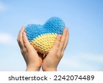 knitted heart of yellow and blue colors of Ukrainian flag in hands of child against sky. Symbol of love for Ukraine, patriotism. Support Ukraine. Stop war. Place for text. Patriotic background.