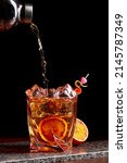 Small photo of Alcohol cocktail pouring from shaker into gless with orange peel ice on stone desk. black background