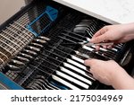 Small photo of Person unloading open dishwasher machine with clean stainless cutlery set: knives, spoons and forks. Inside dishwashing machine with cutlery tray