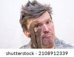 Small photo of Portrait of a geezer holding a dusty pipe wrench in front of his face. Unshaven bully threatening with a rusty hand tool. Adult male with ruffled hair. Indoors. Selective focus.