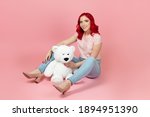 smiling, satisfied woman in jeans with red hair with a large white teddy bear sitting on the floor , isolated on a pink background.