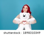 close-up a smiling woman in a white dress with red hair gently hugs a white teddy bear and makes a heart symbol from her fingers isolated on a blue background.