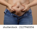 Small photo of Woman squeezing showing fat sagging tummy in blue unzipped jeans on beige background. Sudden weight gain. Visceral fat. Body positive. Tight little clothes. Need for wardrobe change.