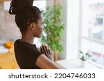 Rearview mindful and meditating young multicultural woman in black sportswear practicing yoga exercises, standing and keeping hands in namaste mudra position. Fitness at home, looking out window