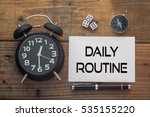 Daily Routine written on paper with wooden background desk,clock,dice,compass and pen.Top view conceptual
