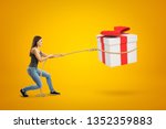 Side view of young woman standing with bent knees and pulling big gift box in air which she has lassoed, on yellow background. Bargain bin. Good eye for bargain. Get what you want.