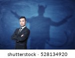 Small photo of A businessman standing with his hands across and his shadow on a blue empty blackboard behind him with hands overspread, horns and a tail like a devil has. Self-confident behavior. Hidden feelings