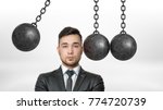 Small photo of A businessman in a front view stands with his head dangerously put between several wrecking balls and one ready to hit. Dangerous position. Put in jeopardy. Problems ahead.
