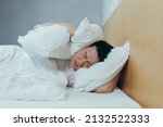 Small photo of A man at home tries to fall asleep, noisy neighbors interfere with sleep, an Asian closes his ears with pillows, tired after work
