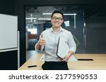 Small photo of Portrait of successful asian teacher, man in modern classroom, looking at camera and smiling, wearing glasses holding laptop, showing thumbs up gesture affirmative