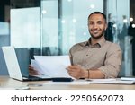 Portrait of young successful hispanic businessman inside office, man smiling and looking at camera, paper worker happy with achievement results sitting at workplace with laptop.