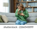 Small photo of Senior beautiful woman sitting sick on sofa at home. He coughs, covers his mouth with his hand, holds his chest. Feels pain, suffers from asthma, allergies, flu, cold.